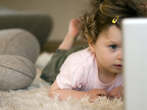 Una niña recostada sobre una alfombra, frente a una laptop.