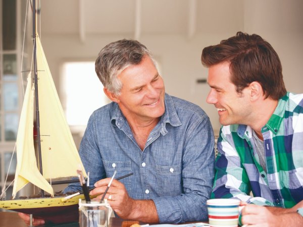 Dos hombres conversan en una mesa, mientras pintan un barco en miniatura.